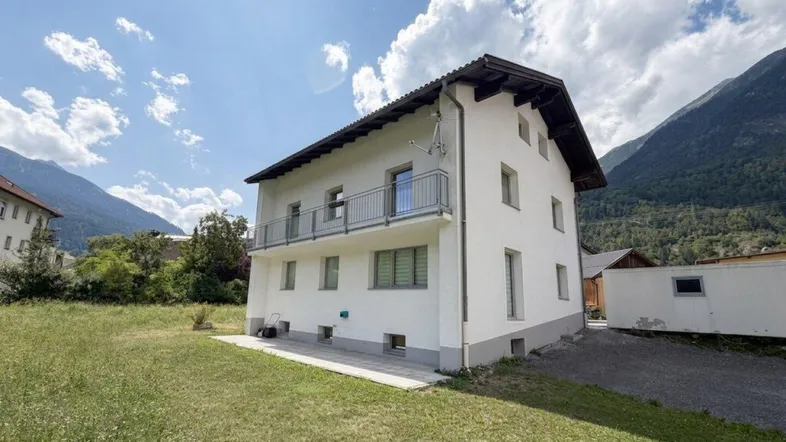 Weißes Wohnhaus mit Balkon und Bergblick, umgeben von grüner Landschaft.