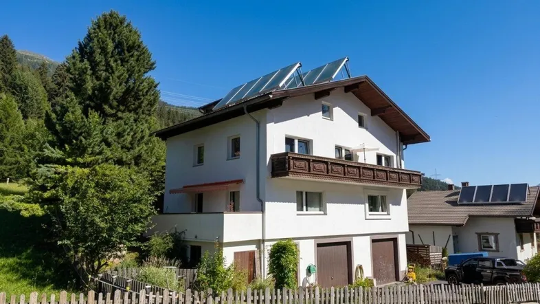 Weißes Wohnhaus mit Holzbalkon, Solaranlage auf dem Dach und umzäuntem Vorgarten.