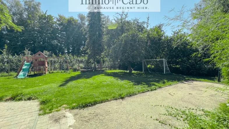 Großer Garten mit Rasenfläche, Kinderspielplatz, Fußballtor und altem Baumbestand unter blauem Himmel.