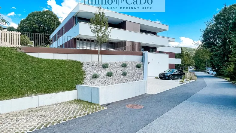 Modernes Mehrfamilienhaus mit mehreren Balkonen, umgeben von Grünflächen und einer Zufahrt.