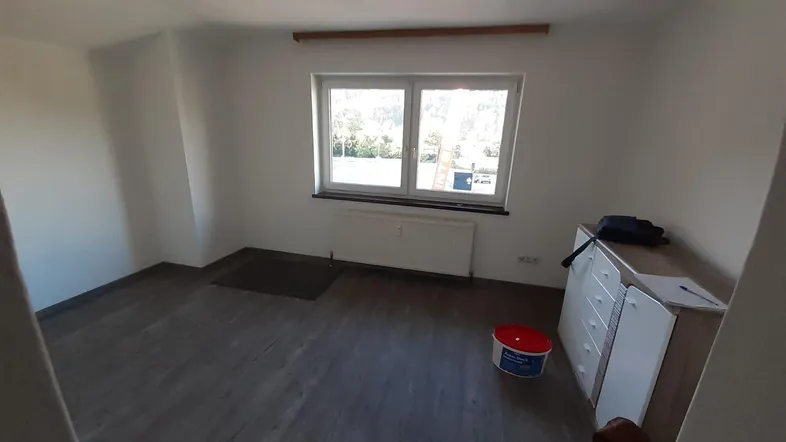 Helles, leeres Zimmer mit Laminatboden und Fenster, das auf die Umgebung blickt.