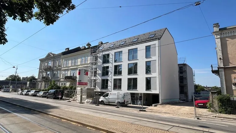 Neubaugebäude im Bau mit Gerüst an der Fassade, umgeben von städtischer Umgebung und Straßenbahnschienen.