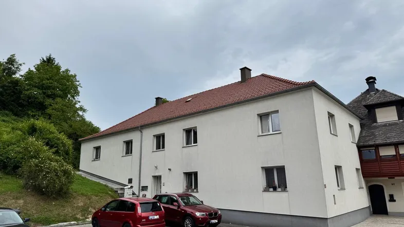 Weißes Mehrfamilienhaus mit rotem Ziegeldach und Parkplätzen vor einem grünen Hügel.