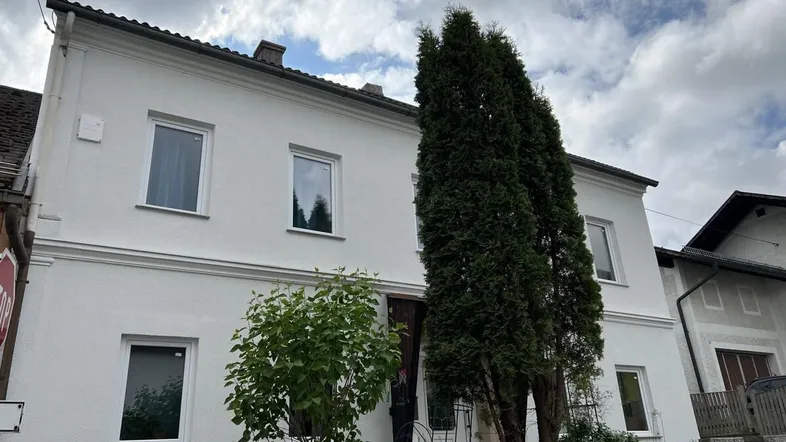 Weißes Wohnhaus mit traditionellem Dach und hohen Bäumen vor der Fassade unter bewölktem Himmel.
