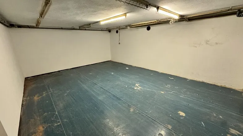 Geräumiger Kellerraum mit blaugrauem Boden und sichtbaren Leitungen an der Decke, beleuchtet von Neonröhren.