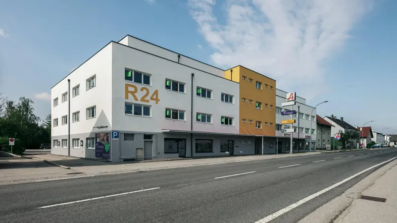 Modernes Gebäude mit weißer und gelber Fassade, Geschäften im Erdgeschoss und Parkplätzen davor.