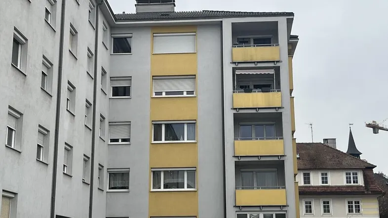 Mehrstöckiges Wohngebäude mit grauer Fassade und gelben Balkonen unter bewölktem Himmel.