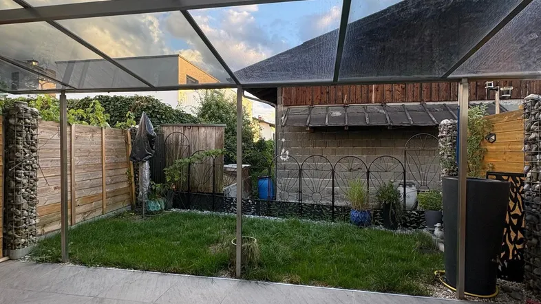 Überdachte Terrasse mit modernen grauen Fliesen und angrenzendem kleinen, eingezäunten Gartenbereich.
