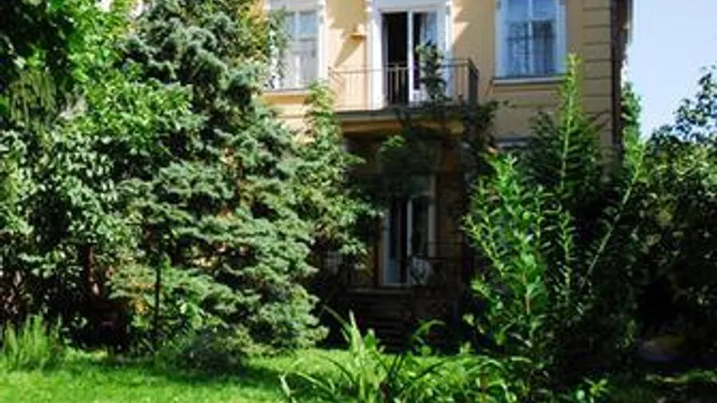 Gelbes Gebäude mit rotem Dach, umgeben von üppigem Grün und einem gepflegten Garten.