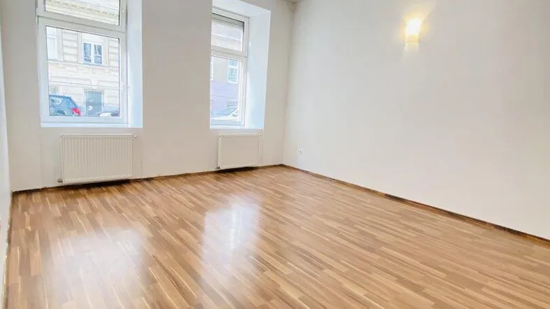 Helles, leeres Zimmer mit Laminatboden und zwei Fenstern, die auf die Straße blicken.
