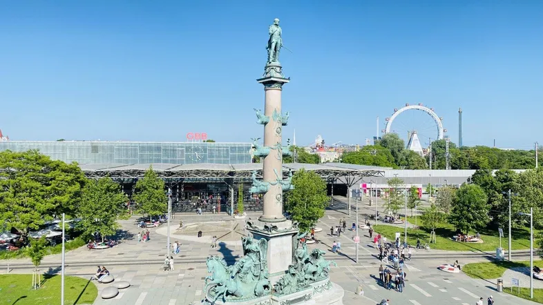 Panoramablick über einen belebten Stadtplatz mit Denkmal und Riesenrad im Hintergrund.