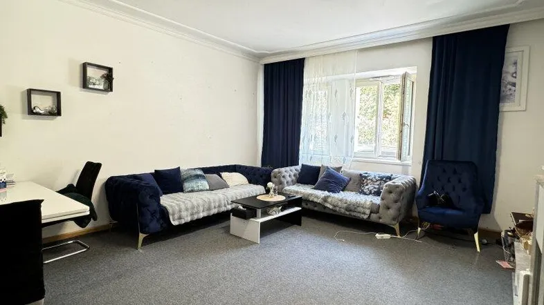Geräumiges Wohnzimmer mit grauem Teppichboden, blauen Sofas und einem Arbeitsbereich am Fenster.