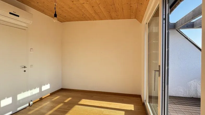 Helles Zimmer mit Holzbalkendecke, Parkettboden und Zugang zu einem Außenbereich.