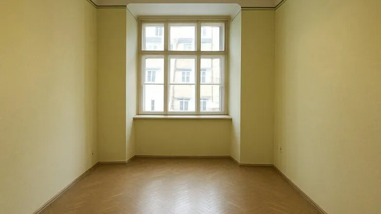 Helles Zimmer mit Fischgrätparkettboden und einem großen Fenster, das viel Tageslicht hereinlässt.