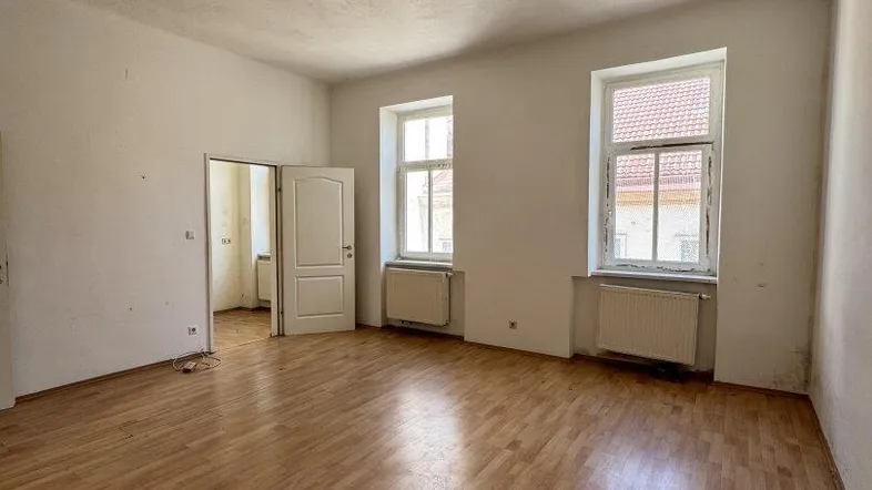 Geräumiges, leeres Wohnzimmer mit Holzboden, hohen Decken und zwei Fenstern, die renovierungsbedürftig sind.