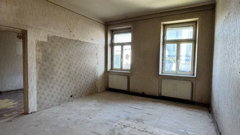 Leerer Raum in Renovierungszustand mit abgelöster Tapete und Blick aus zwei Fenstern.