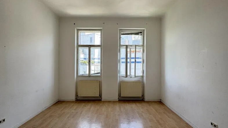 Helles Zimmer mit Holzboden und zwei Fenstern, bereit zur individuellen Gestaltung.