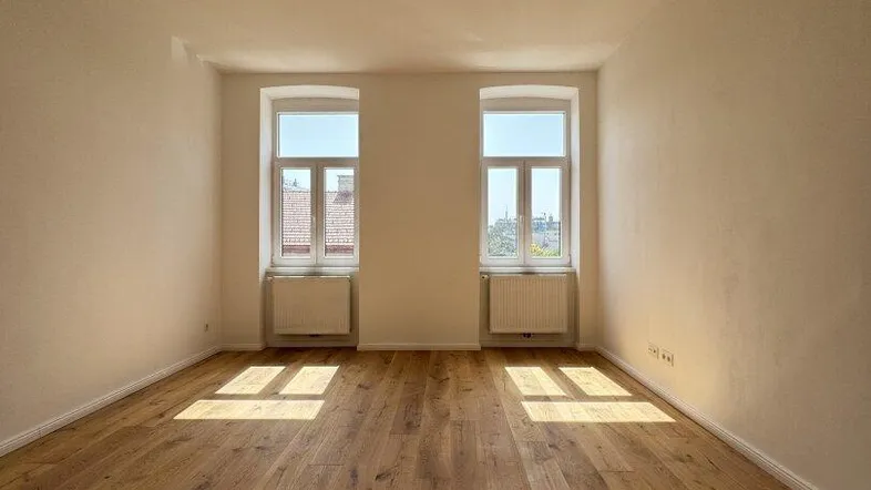 Helles, leeres Zimmer mit Holzboden und zwei Fenstern, durch die Sonnenlicht einfällt.