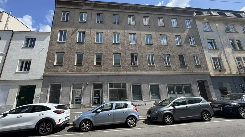 Graues Mehrfamilienhaus mit mehreren Etagen und Geschäften im Erdgeschoss, geparkt an einer Straße.