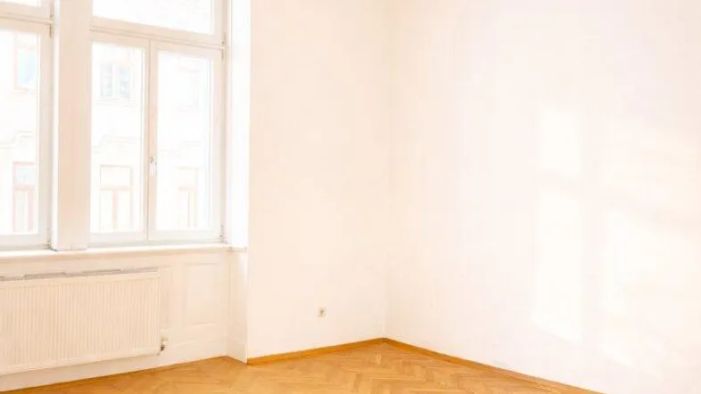 Helles, leeres Zimmer mit Fischgrätparkettboden und großen Fenstern, durch die Sonnenlicht fällt.