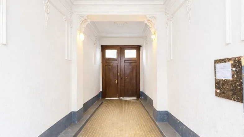Langer, heller Flur mit historischen Fliesen, Stuckelementen an der Decke und einer Holztür am Ende.