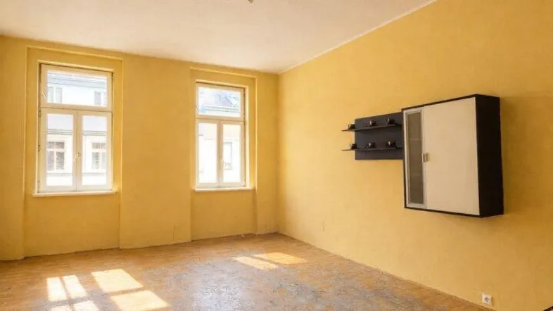 Helles Zimmer mit gelben Wänden, zwei Fenstern und abgenutztem Parkettboden.