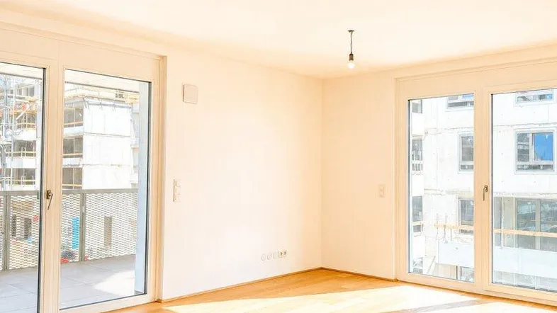 Helles Zimmer mit Holzboden und großen Fenstern, die viel Tageslicht hereinlassen und Zugang zum Balkon bieten.