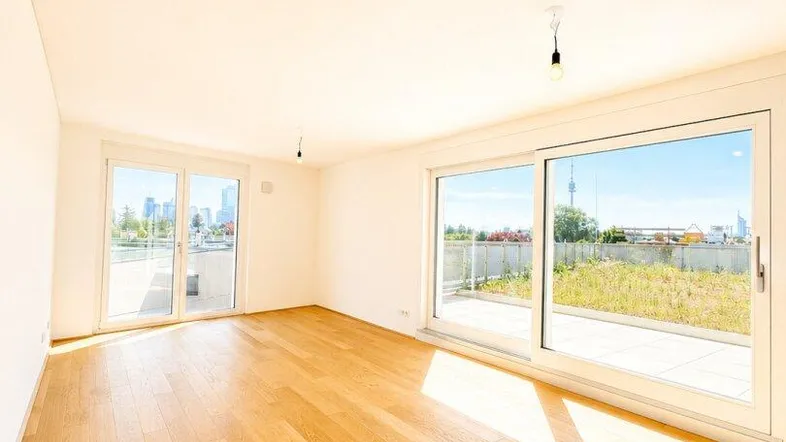 Helles, leeres Zimmer mit Holzboden und großen Schiebetüren zu einer Terrasse.