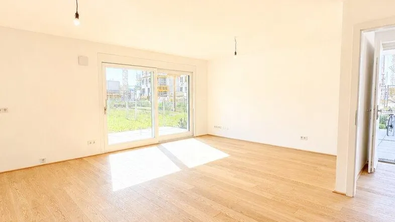 Helles, leeres Wohnzimmer mit Holzboden und Zugang zu einem kleinen Gartenbereich.