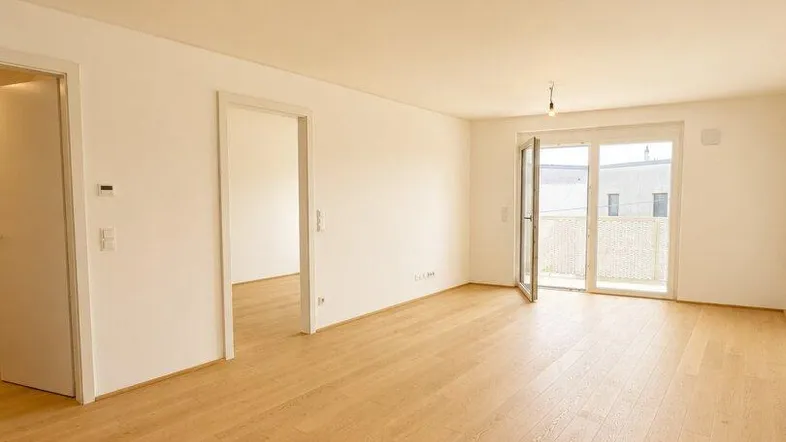 Helles, leeres Wohnzimmer mit Holzboden und Zugang zu einem Balkon oder einer Terrasse.