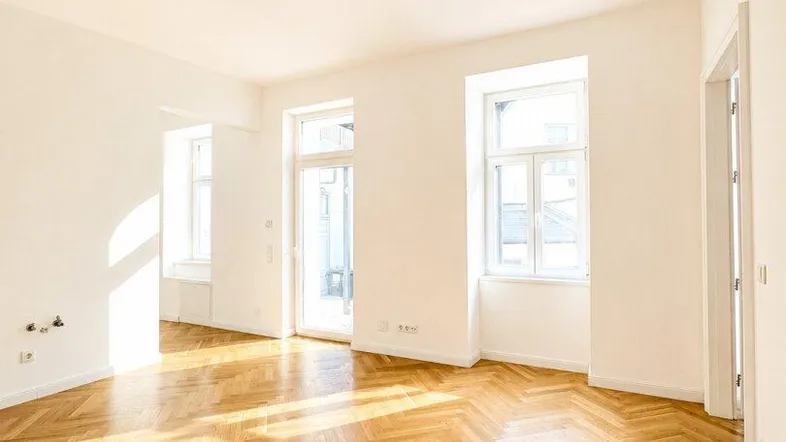 Helles, leeres Zimmer mit Fischgrätparkett und großen Fenstern, das viel Sonnenlicht hereinlässt.