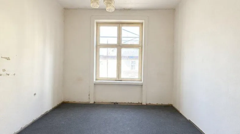 Leerer Raum mit dunklem Teppichboden, einem Fenster und einer einfachen Deckenleuchte, bereit zur Renovierung.