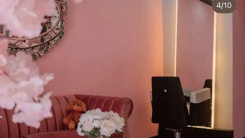 Rosa gestrichener Raum mit Samtsofa, beleuchtetem Spiegel und Friseurstuhl, dekoriert mit Blumen und einem Teddybär.