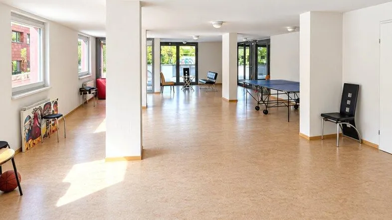 Großer, heller Gemeinschaftsraum mit Korkboden, Säulen und einem Tischtennistisch im Hintergrund.