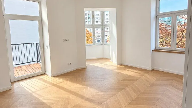 Helles, leeres Zimmer mit Fischgrätparkett, großen Fenstern und Zugang zu einem französischen Balkon.