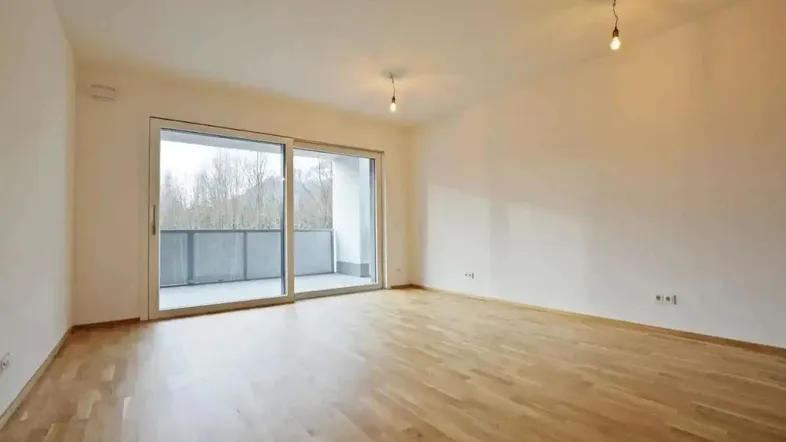 Helles, leeres Zimmer mit Holzboden und Zugang zu einem Balkon durch eine große Glasschiebetür.