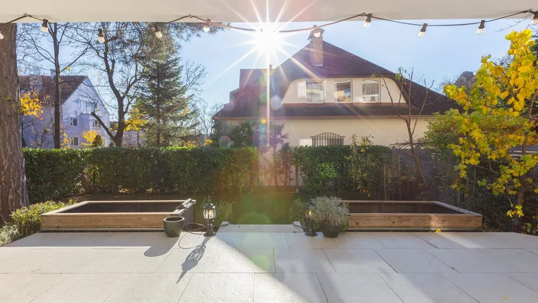 Sonnige Terrasse mit hellen Fliesen, erhöhten Pflanzenbeeten und Blick auf das Nachbargebäude.
