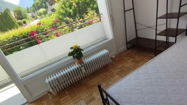 Helles Schlafzimmer mit Parkettboden, einem Regal und Zugang zu einem Balkon mit grüner Aussicht.