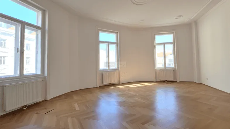 Geräumiges Wohnzimmer mit Fischgrätparkett, hohen Decken und großen Fenstern, die viel Tageslicht hereinlassen.