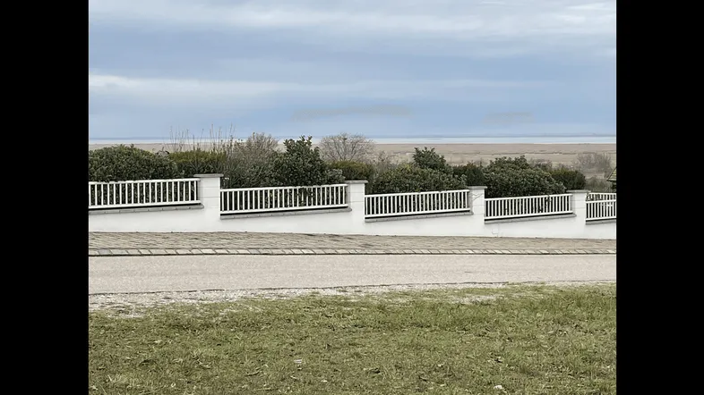 Blick über einen weißen Zaun auf eine weite Wasserfläche und Schilflandschaft unter bewölktem Himmel.