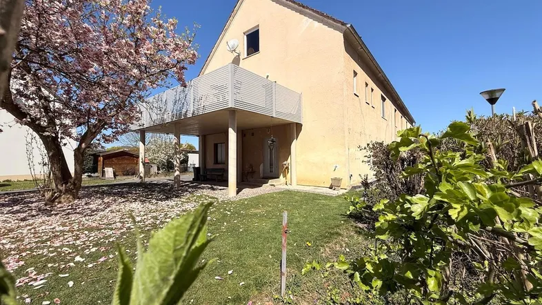 Hausfassade mit blühendem Baum, Garten und Balkon unter blauem Himmel.