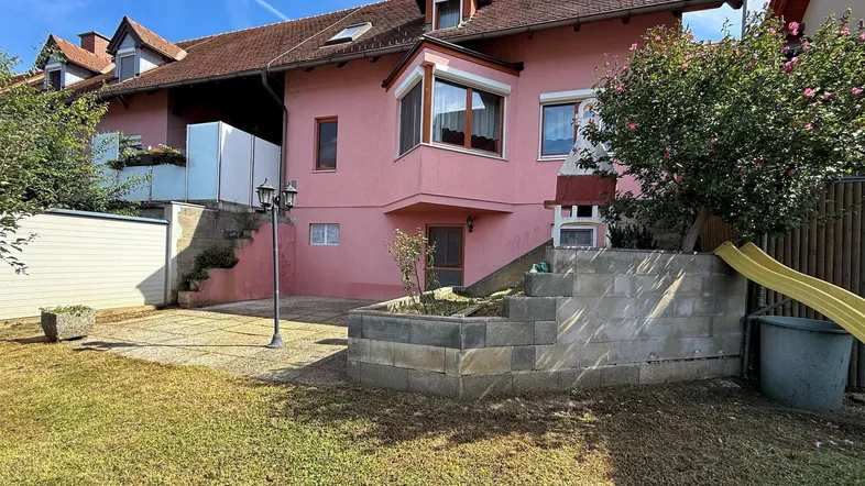 Hinterhof mit Rasenfläche, Kinderrutsche und gemauertem Grillbereich, umgeben von einem rosa Haus.