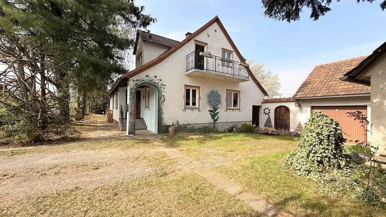 Ansicht des Hauses mit kleinem Balkon, Vorgarten und Zufahrt unter Bäumen.