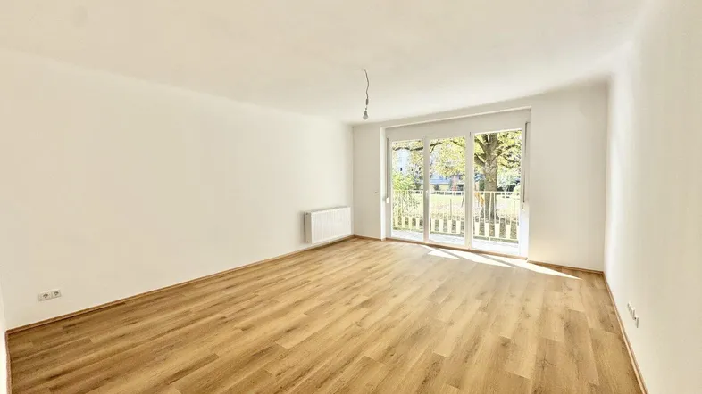 Helles, leeres Zimmer mit Laminatboden und Zugang zu einem Balkon oder Gartenbereich.