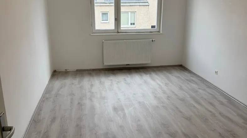 Helles, leeres Zimmer mit Laminatboden und Fensterblick auf umliegende Gebäude.