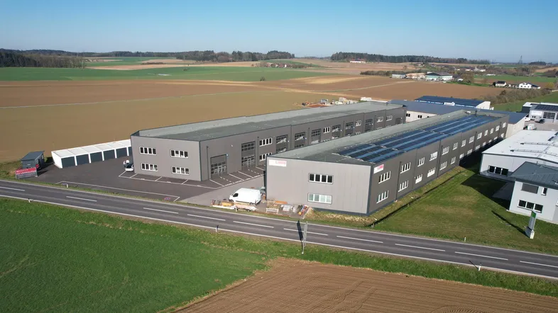 Luftaufnahme eines modernen Gewerbegebäudes mit Photovoltaikanlage auf dem Dach, umgeben von Feldern und einer Straße.