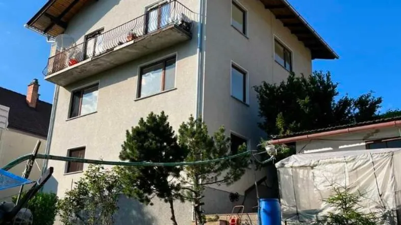 Mehrstöckiges Wohnhaus mit Satteldach und Balkonen, umgeben von einem grünen Garten.