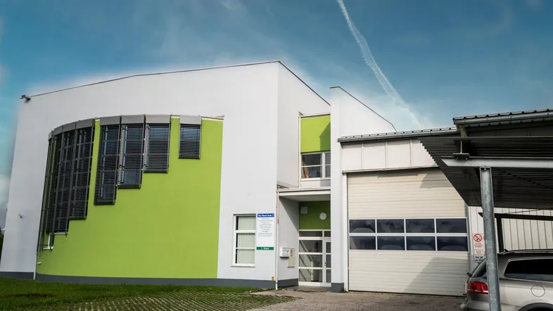 Modernes Gebäude mit weißer und hellgrüner Fassade, großen Fenstern und einem Carport auf der rechten Seite.