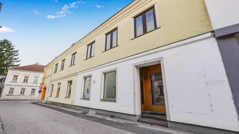 Die gelbe Fassade eines älteren Gebäudes mit mehreren Fenstern und einem einladenden Holzeingang ist zu sehen.