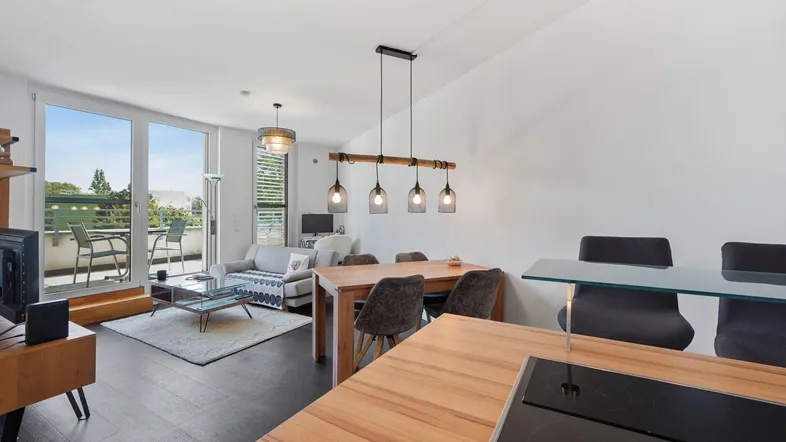 Heller offener Wohn-Essbereich mit moderner Küche, Holzboden und Zugang zum Balkon.